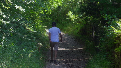 Obraz premium Older man hiking alone on forest path, walking uphill in shorts and white shirt, surrounded by dense greenery and quiet wooded atmosphere