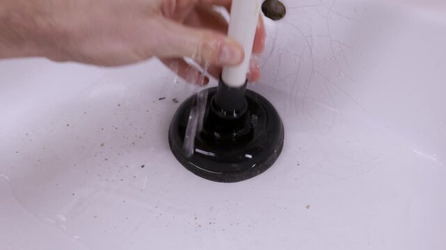 A hand uses a plunger to clear a blockage in a sink filled with dirty water. The water drains away leaving hair and grime. This illustrates plumbing maintenance.