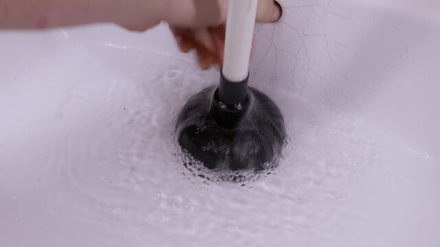 A male hand uses a black plunger to clear a blockage in a white ceramic sink filled with water. The basin has visible cracks on the surface. This depicts a concept of DIY plumbing repair.