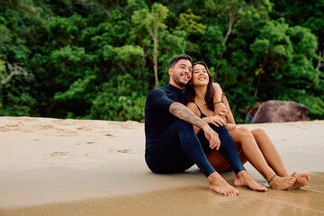 Fototapeta premium Smiling young couple in wetsuits sitting on a tropical beach, looking away while enjoying their summer holiday