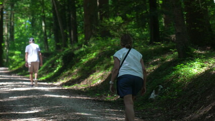 Obraz premium Older couple walking on shaded forest path, both wearing casual clothing and moving uphill along dappled gravel trail in summer woods