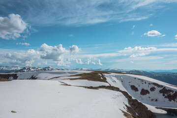 Scenic landscape with sunlit snowfield against snow cornice and big snowbound mountain range far away under dramatic cloudy sky. Vast view with snowy glade in high mountains under clouds in blue sky. © Daniil