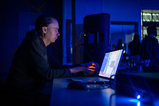 A man works as a sound engineer or DJ in a dark setting, using a laptop and mixing console to adjust audio levels for a concert, club, or disco.