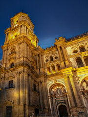 A grand historic Malaga Cathedral bathed in warm golden light, highlighting intricate stonework and a bell tower against a deep blue evening sky, Malaga, Spain
