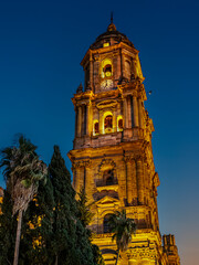 A grand historic Malaga Cathedral bathed in warm golden light, highlighting intricate stonework and a bell tower against a deep blue evening sky, Malaga, Spain