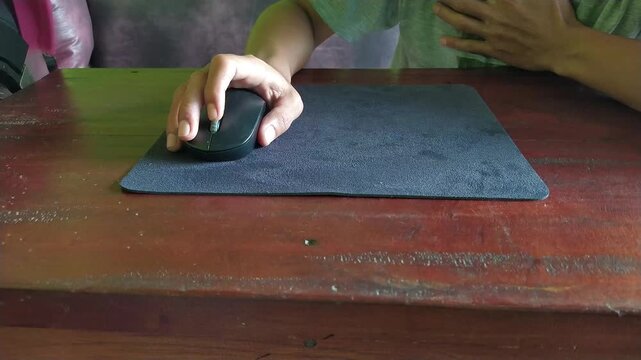 Close up of a hand operating a computer mouse on a mousepad over a wooden desk. Concept of work, browsing, online activity, productivity, or gaming.
