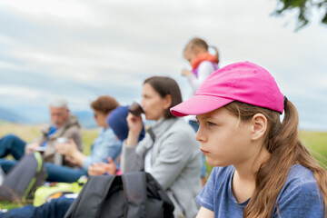 Pensive Young Girl in Pink Cap Portrait during Family Picnic Gathering on Grassy Meadow with Blurred Multi-Generational Relatives