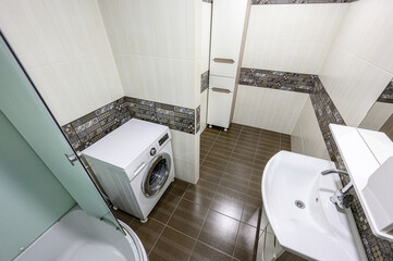 A functional bathroom with a glass-enclosed shower, white sink, wall-mounted cabinet, washing machine, dark tiled floor, and decorative tile border; clean and well-organized