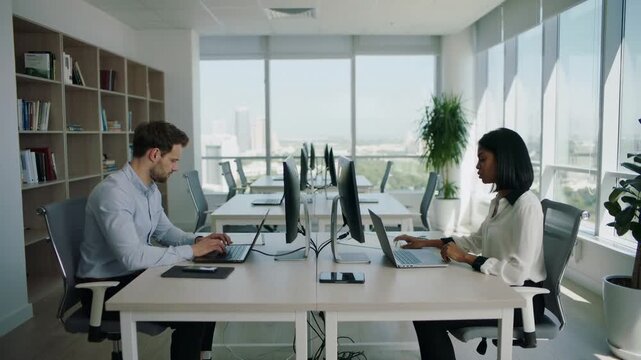 Caucasian male and African female professionals working on laptops at modern office desks, with large windows and greenery providing a bright, collaborative workspace
