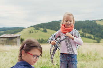 Joyful Toddler Girl in Red Glasses and Orange Plaid Scarf Plays on Grassy Hill with Hillside Barn and Mountains Background