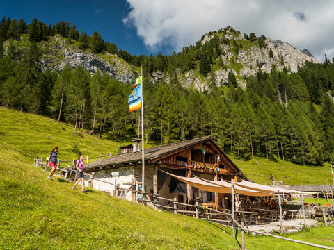 Two hikers approach a mountain hut with outdoor seating, set against a backdrop of forested slopes and rocky peaks. Tartoi hut,Forni di Sopra,Dolomites,Italy