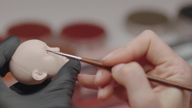 Closeup of skilled artist using brush to paint delicate facial features on handcrafted doll head with focus on precision and artistry