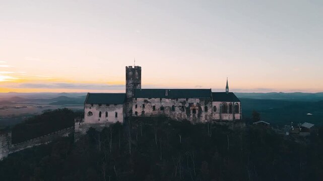 The sun sets behind Bezdez Castle. The sky changes colors as the evening approaches. The castle stands tall on the hill overlooking the landscape.