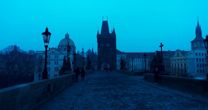 Quiet winter morning on Charles Bridge in Prague with only a few people crossing. Cold blue atmosphere and soft dawn light create a peaceful and cinematic city scene.