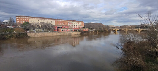 Obraz premium Paisaje urbano junto al río con edificio de ladrillo y puente de piedra antiguo bajo cielo nublado en Logroño, España