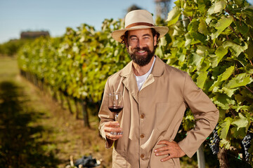 A vineyard owner stands among lush grapevines, holding a glass of red wine. The sun shines down on this family-run wine production site, highlighting the rich harvest. © Stockphotodirectors