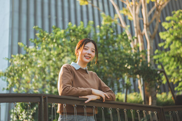 Smiling Asian woman leaning on terrace railing with modern building and green trees background. Urban lifestyle portrait of happy young female enjoying morning sunlight.