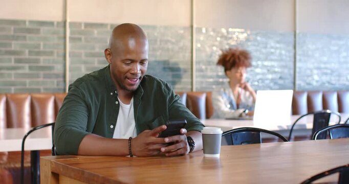 African American man in green top reading smiling scrolling phone lifting cup; woman typing laptop