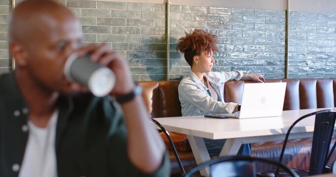 Two African American patrons sitting at cafe, man sipping cup, woman leaning back, using laptop