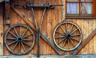 Obraz premium Two old wagon wheels are mounted on a wooden wall beside a small window. The wood exhibits a rich, weathered texture and rustic charm.