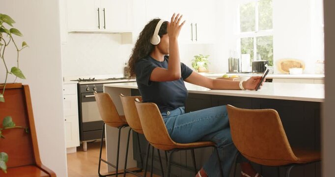 African American woman sitting at kitchen island answering phone alert, adjusting hair, headphones