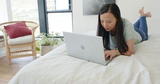 Woman lying on stomach on bed, typing on silver laptop, working to finish report