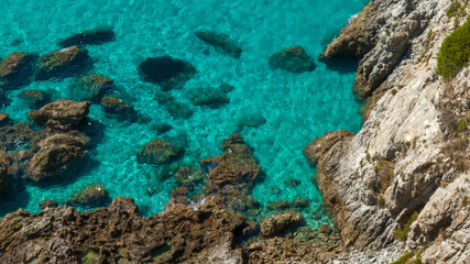 Obraz premium Aerial close-up of crystal-clear turquoise water meeting a rocky shoreline. Large brown rocks are visible beneath the surface, highlighting the incredible transparency of the Mediterranean sea.