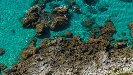 Obraz premium Aerial close-up of crystal-clear turquoise water meeting a rocky shoreline. Large brown rocks are visible beneath the surface, highlighting the incredible transparency of the Mediterranean sea.