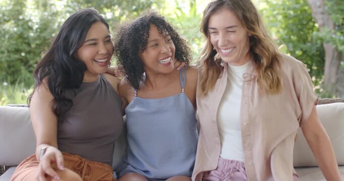 Diverse female friends adjusting scooting inward on outdoor sofa cushions hugging posing for camera