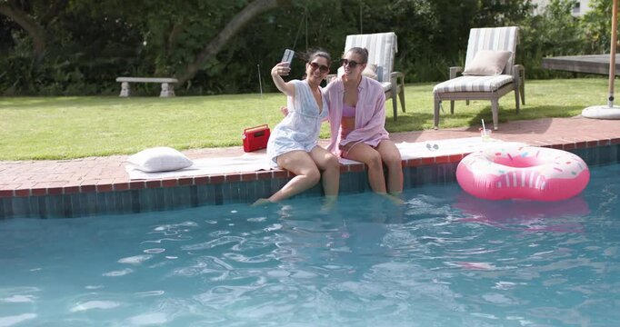 Two female friends sitting on pool edge, one raising smartphone, capturing selfies near pink float
