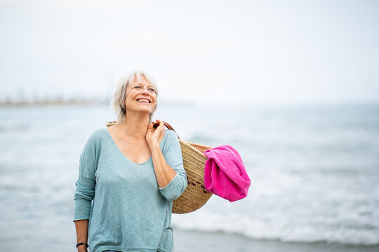 Joyful senior woman standing at the shore with beach bag