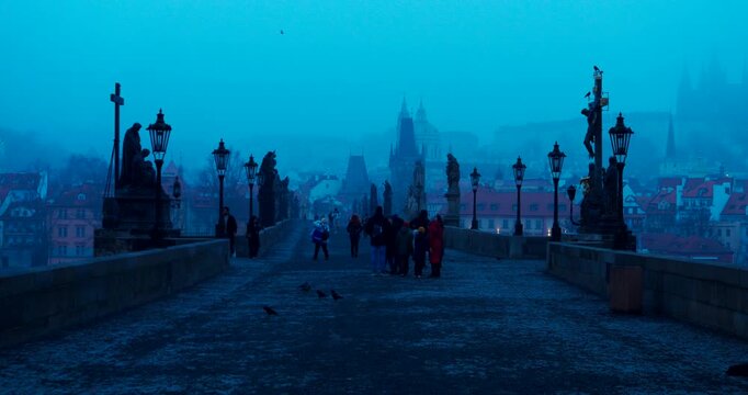 Quiet winter morning on Charles Bridge in Prague with only a few people crossing. Cold blue atmosphere and soft dawn light create a peaceful and cinematic city scene.