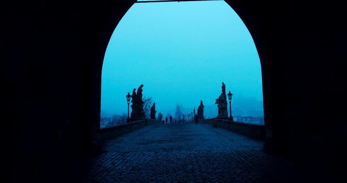 Quiet winter morning on Charles Bridge in Prague with only a few people crossing. Cold blue atmosphere and soft dawn light create a peaceful and cinematic city scene.