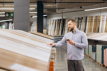 Man shopping for wood laminate flooring, comparing samples while holding a smartphone in a hardware...