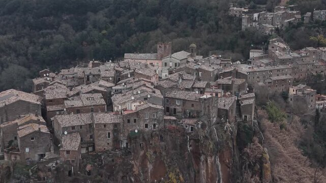 Aerial view of Calcata Vecchia, an ancient medieval village perched on a rocky cliff in Lazio, during a gloomy day. Historic stone houses surrounded by forest and dramatic landscape of a scenic view.
