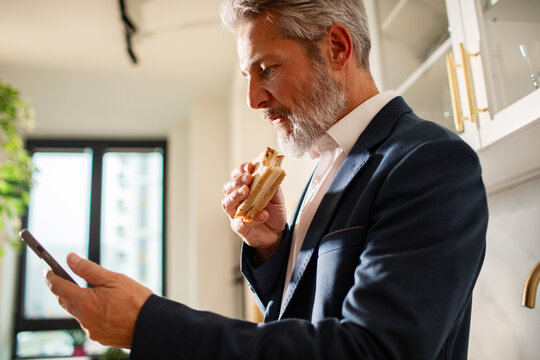 Mature businessman eating a sandwich while using smartphone in home kitchen