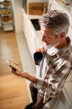 Middle-aged man drinking coffee and using smartphone in home kitchen