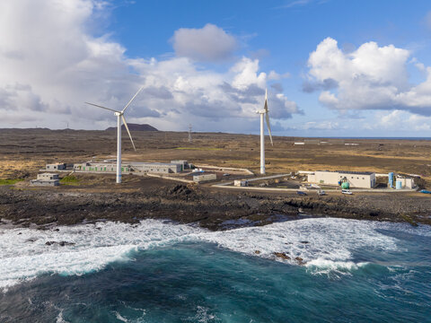 Aerial view of wind turbines standing tall against the rugged, dark coastline where turquoise waves crash, contrasting with the industrial buildings, Corralejo, Fuerteventura, Spain.
