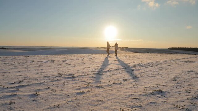 Active married couple walks in snowy meadow in winter evening. Romantic man and woman date joining hands on white valley. Tourists in love enjoy promenade at back sunset light