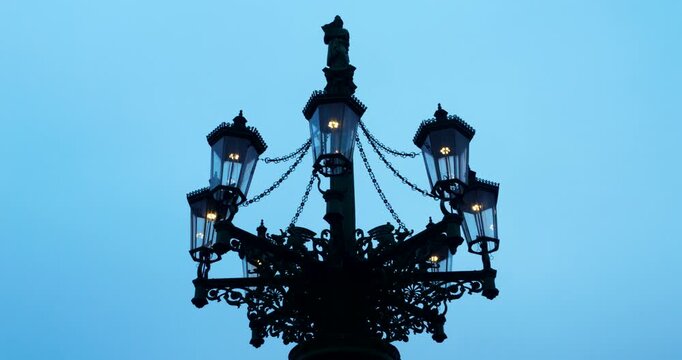 Close-up of historic iron street lamps glowing in Prague&rsquo;s Old Town during a dark blue winter morning. Warm city lights contrast with the cold silhouette skyline in a moody atmosphere.