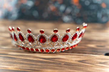 Ornate silver tiara adorned with red gemstones placed on a wooden surface, showcasing intricate design and sparkling details against a blurred colorful background © Андрей Знаменский