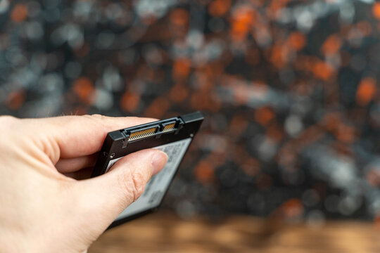 Hand holding a solid-state drive (SSD) with connectors visible, against a blurred background featuring a textured surface with orange and black patterns