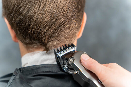 Male hairdresser using electric clippers to trim the back of a client's hair while wearing a black cape in a modern barbershop setting