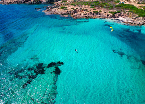Aerial view of a kitesurfer gliding across the shimmering turquoise waters near a rugged, rocky coastline, Corse, Corse, France.