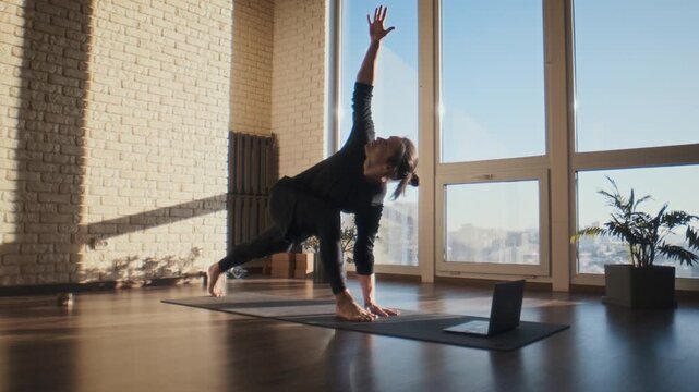 Focused man performs yoga twist near laptop in bright studio