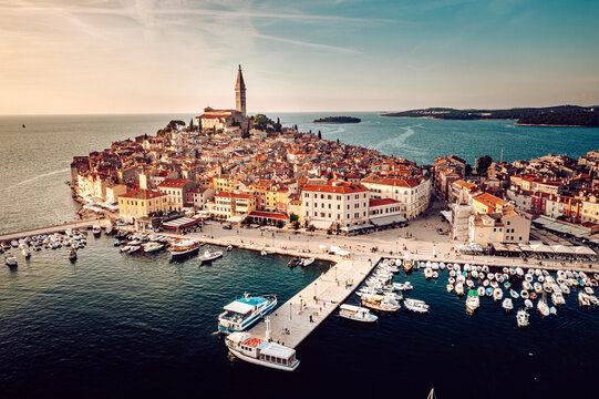 Aerial view of terracotta rooftops cascade down to the azure harbor, where boats bob gently beneath the watchful gaze of St. Euphemia's bell tower, Rovinj, Istria County, Croatia.