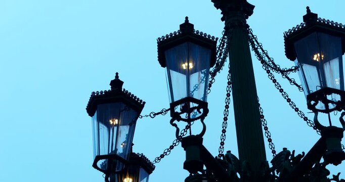Close-up of historic iron street lamps glowing in Prague&rsquo;s Old Town during a dark blue winter morning. Warm city lights contrast with the cold silhouette skyline in a moody atmosphere.