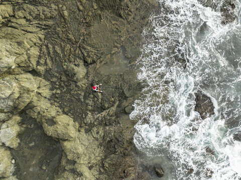 Aerial view of the rugged coastline meets the foamy waves, a solitary angler in red amidst the rocks, Siung Beach, Gunungkidul Regency, Special Region of Yogyakarta, Indonesia.