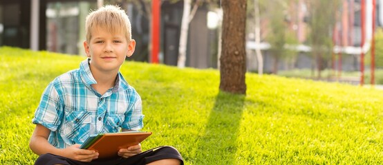 Happy child sitting on the field holding tablet. Boy sitting on the grass on sunny day. Home schooling or playing a tablet