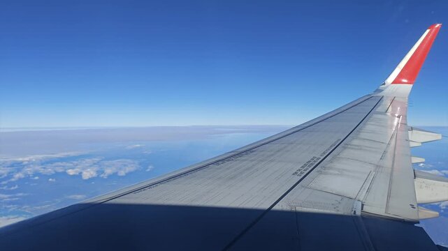 Aerial view of an airplane wing with a red tip flying high above clouds in a clear blue sky.